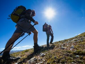 Due escursionisti con zaini che percorrono percorso in salita, uno dei due ha i bastoni, cielo azzurro e sole