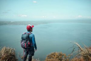 Ragazzo con zaino e berretto che guarda il mare dall'alto mentre si trova su un sentiero