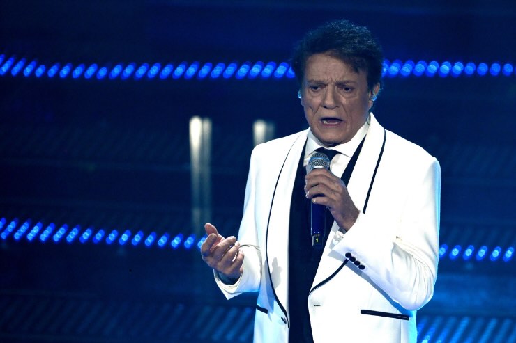 Massimo Ranieri vestito di bianco che canta sul palco blu