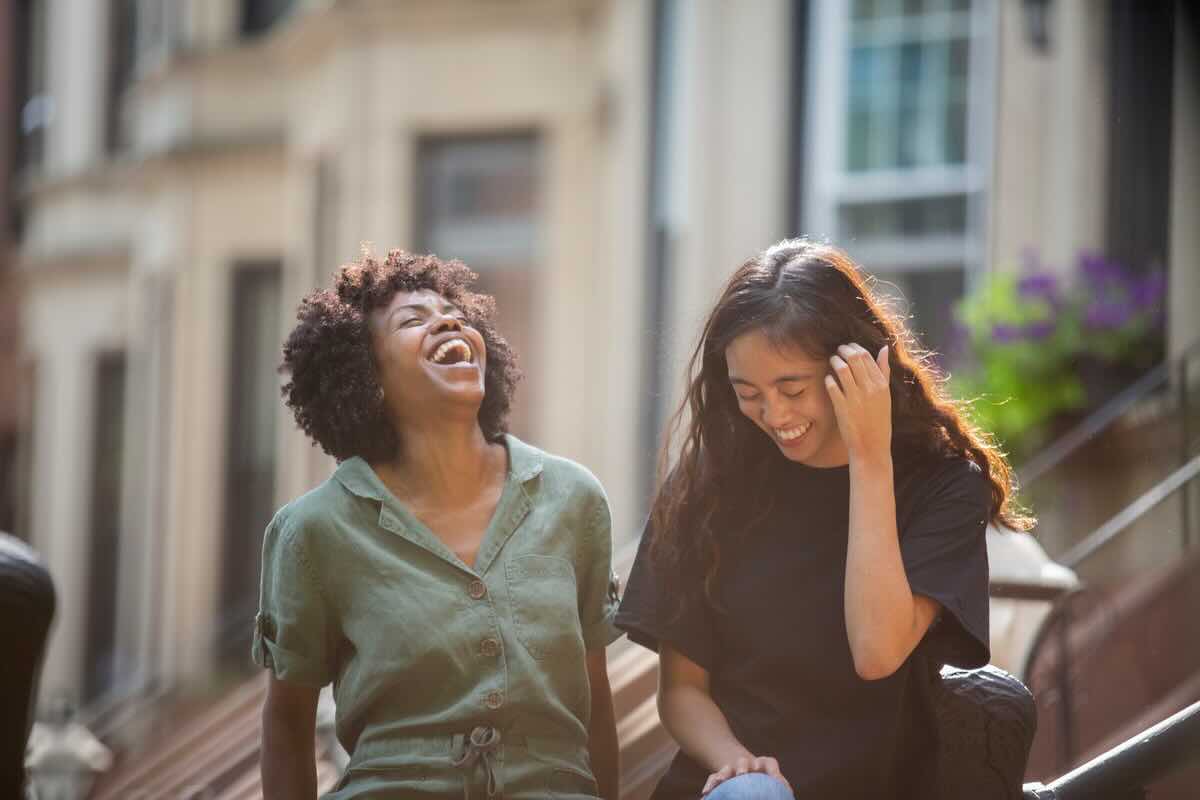 Due ragazze di colore che ridono di gusto, una delle due si tocca i capelli