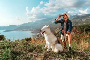 Uomo che guarda con il cannocchiale e cane bianco al suo fianco, immersi nella natura con mare sullo sfondo